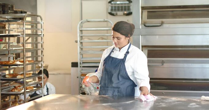 Diverse female bakers at bakery in coats using spray bottle, cleaning table and handling rack trays