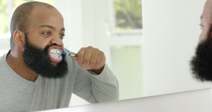 Bearded man starting routine, standing by mirror brushing teeth with blue-yellow brush, making foam