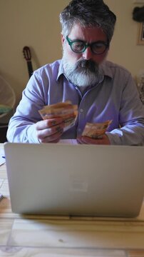 Middle-aged man with a beard sitting at his home desk, anxiously counting euro banknotes while managing his financial budget on a laptop, showing stress and concern about his expenses