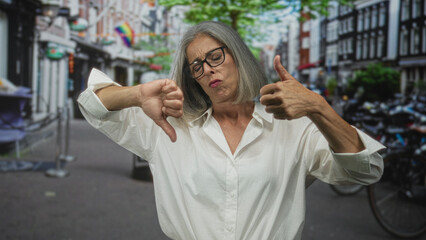 Fototapeta premium Middle aged woman smiling and showing thumbs up and thumbs down on a busy city street lined with bicycles and brick buildings; ambivalence.