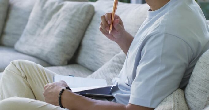 Asian man reaching for rose-gold pen writing in notebook pausing and planning on sofa, copy space