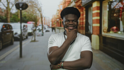Fototapeta premium Man with hand on chin and open mouth on street near storefront, wearing white t shirt and beaded necklace; playful confidence.