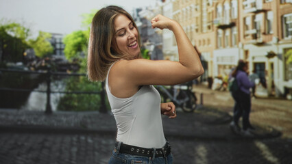 Fototapeta premium Woman flexes bicep and smiles in white tank top beside canal on a cobbled street with buildings and bicycle; confidence empowerment.