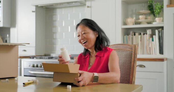 Senior Asian woman wearing red blouse opening box at kitchen table, inspecting white bottle label