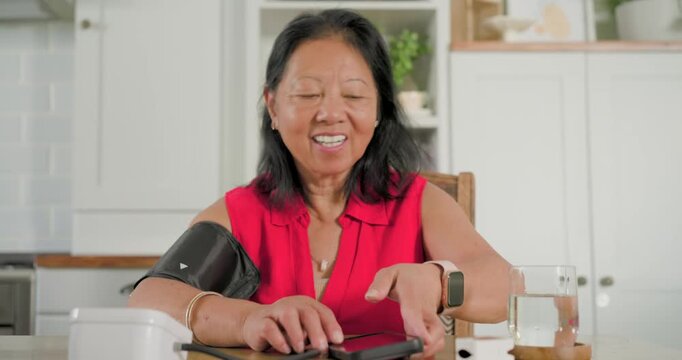 Senior Asian woman in red blouse tapping smartphone, checking BP for health, stowing cuff on base