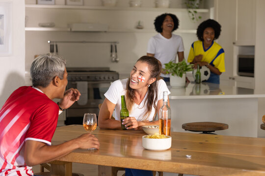 Diverse fans cheering after goal during match in kitchen, woman with red cross holding green beer