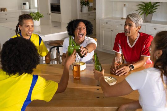 Group of friends in yellow and red jerseys clinking green beer bottles at wooden kitchen island
