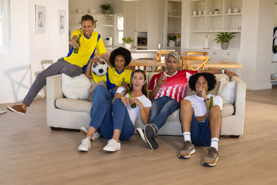 Diverse friends watching soccer match in living room wearing jerseys holding ball and green beers