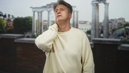 Fototapeta premium Man senior grey hair holds neck with hand showing neck pain on a terrace by ancient building ruins and roman columns; fatigue.