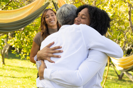 Diverse friends hugging in backyard on grassy lawn under draped fabric canopy, third friend smiling
