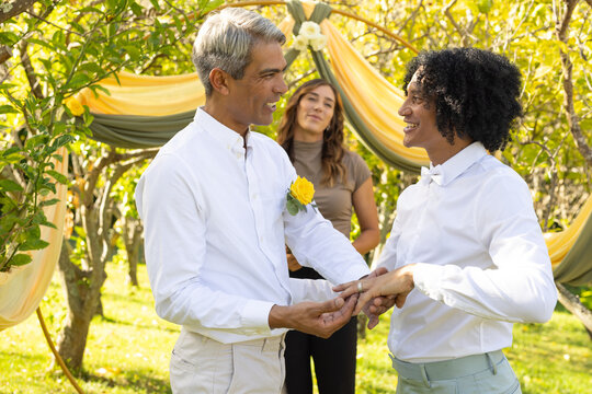 African American men exchanging rings under yellow-green fabric arch in garden, rose pin visible
