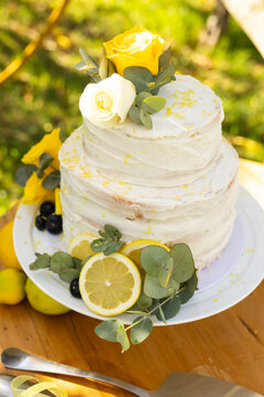 Two-tier buttercream cake sitting on white stand in garden, displaying yellow rose and lemons