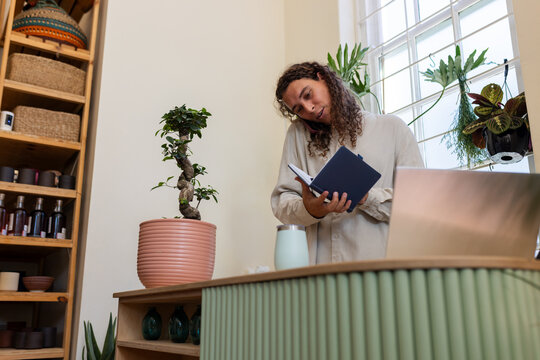 Nonbinary African vendor balancing phone and writing navy notebook in plant shop with laptop