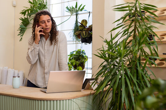 Nonbinary African American in beige top working at curved counter in plant-shop using phone, laptop