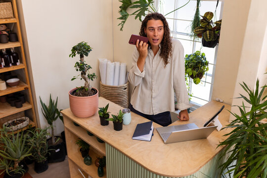 African American non-binary in beige shirt typing on laptop and speaking into phone at plant shop