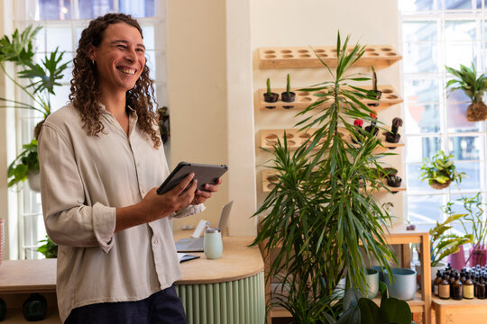Non-binary African American smiling, holding tablet by curved counter in plant shop, copy space