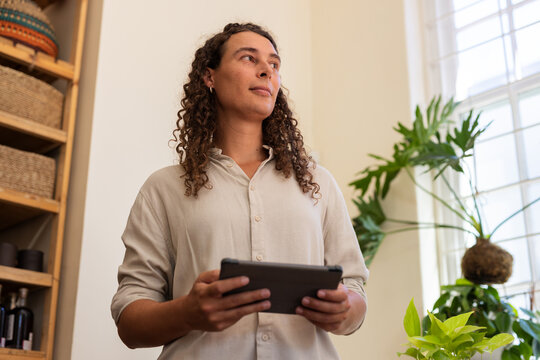 Non-binary person standing, holding tablet, wearing beige shirt, looking upward in home with plants