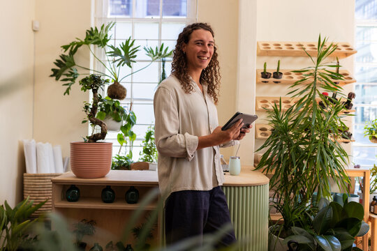 Non-binary African owner standing behind wood counter in plant shop, holding tablet and smartphone