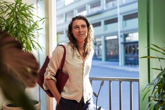 African non-binary person standing in doorway with maroon tote leaning on railing looking to street