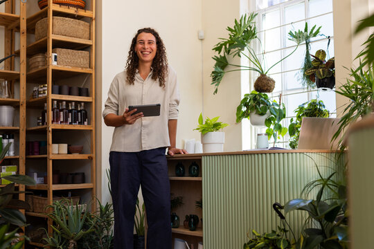 African American non-binary owner standing behind green counter holding tablet with laptop  and  pla