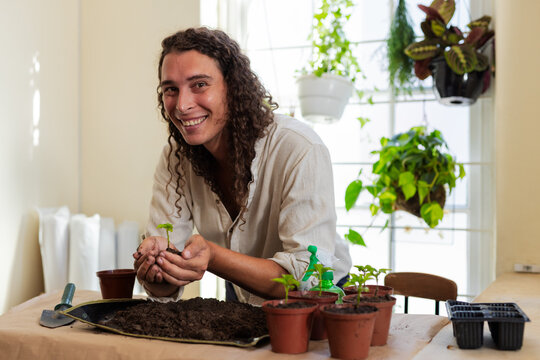 Non-binary person in beige shirt leaning over table at home, holding seedling and trowel