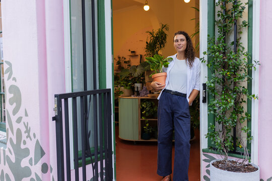 Non-binary person holding terracotta plant in shop doorway wearing white tee navy pants, copy space