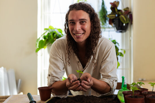 Non-binary adult wearing light shirt with rolled sleeves smiling, potting seedling by window