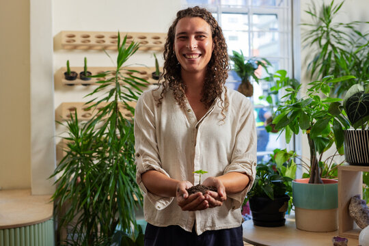 African American nonbinary owner smiling holding seedling in soil at plant shop in beige shirt