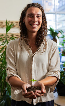 Non-binary person holding small green seedling in dark soil, smiling in plant studio in linen