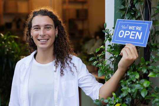 Non-binary person standing at shop entrance holding blue OPEN sign, wearing white shirt, plants