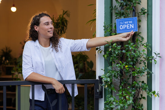 African-American nonbinary owner reaching for blue Yes WE'RE OPEN sign at door amid plants and trim