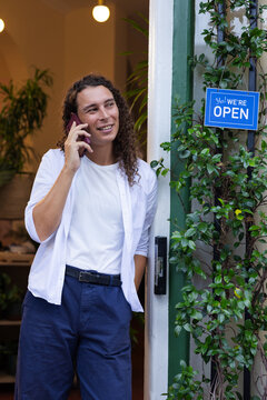 African non-binary shop-owner leaning on green door talking on phone near blue open sign in white-T