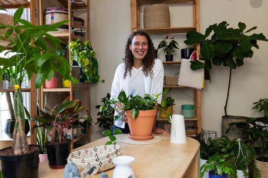 Non-binary person tending terracotta plant pot on wooden counter in plant shop, wearing white shirt