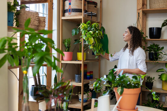 Nonbinary person wearing white shirt tending vine on wood shelf with watering can, copy space