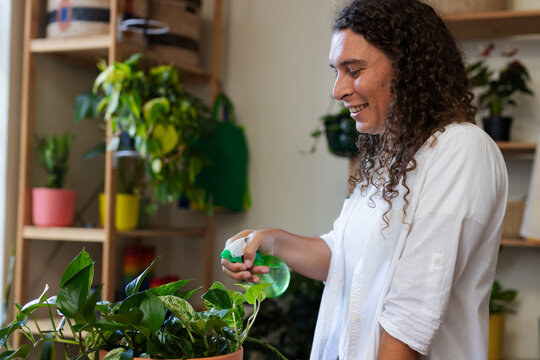 African American nonbinary wearing white shirt misting plant with green spray at home, copy space