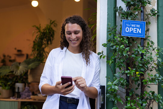 African American nonbinary person standing in shop doorway holding smartphone near open sign