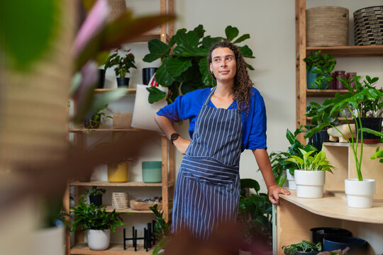 Non-binary proprietor leaning on curved wooden counter at plant shop, wearing striped apron