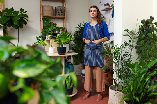 African American non-binary wearing striped apron standing among potted plants at small plant shop