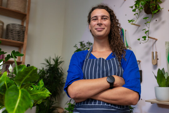 African American non-binary standing arms crossed in plant shop among plants wearing navy apron