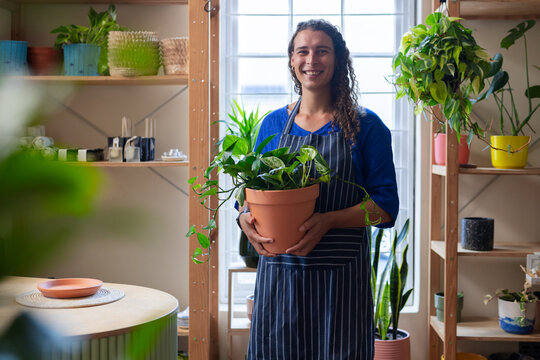 Non-binary shop owner standing in plant shop holding terracotta pothos pot wearing pinstripe apron