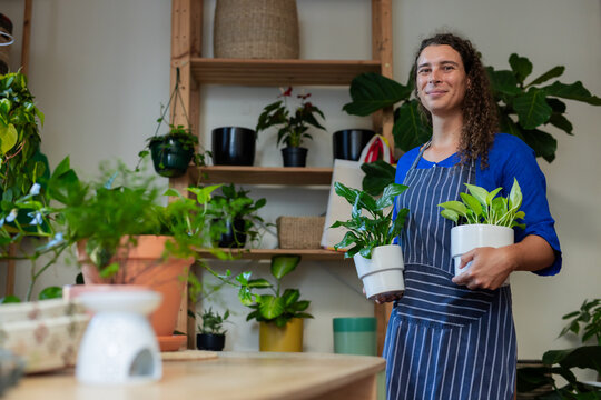 Non-binary person standing inside plant shop wearing striped apron and holding two white pots