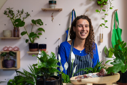 Non-binary person standing at plant shop counter wearing striped apron tending peace lilies