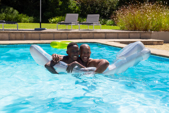 African father and adolescent son leaning on pool float in pool, wearing trunks, neon tube behind