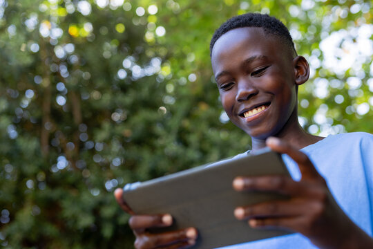 African teen holding tablet in dark case, smiling, looking down at park wearing light blue T-shirt