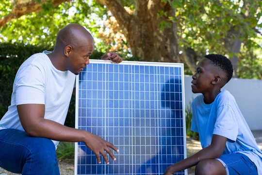 African American man teaching teen male after pointing at blue solar panel while crouching in yard