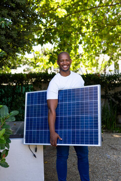 African man wearing white T-shirt and jeans holding solar panel in garden near ledge with cable