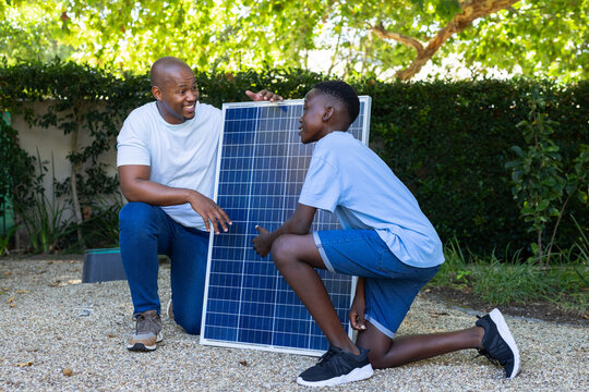 African American father and teenage son kneeling on gravel holding solar panel upright in backyard