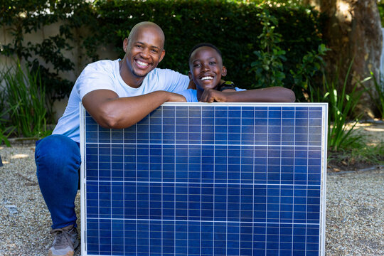 Father and teenage son kneeling behind solar panel on backyard gravel, wearing white tee, smiling