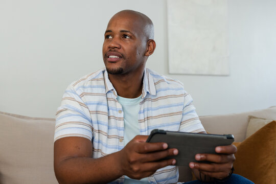 Adult African American man sitting on beige sofa in living room holding tablet, looking up left