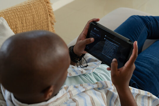 African man reclining on light sofa, holding tablet, wearing wired earbuds and wristband